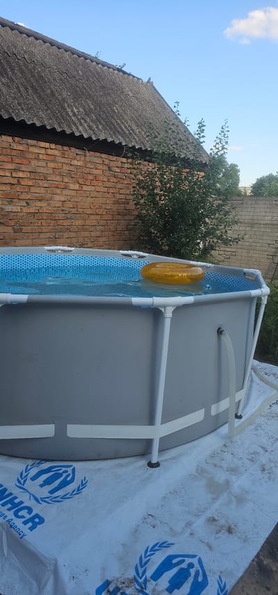 In Berestyn, Ukraine, an above-ground swimming pool is set up outdoors, resting on a white tarp with the UNHCR logo visible. The pool is partially filled with clear water, and a yellow inflatable ring floats on the surface. The pool has a grey exterior and white support poles. Behind the pool, a brick wall with a corrugated roof is visible. To the right of the brick wall, a dense green bush grows. Beyond that, a grey cinder block wall marks the property line, with more greenery visible behind it. The sky above is blue with a few white clouds. The overall scene suggests a residential backyard environment in the city.