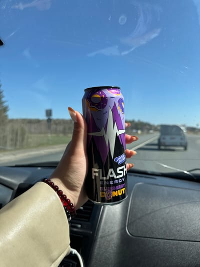 A person is holding a can of Flash Energy Blueberry Donut drink in their hand inside a vehicle. The vehicle appears to be on a road with other vehicles in the distance. Outside the window is a blue sky and some trees. A portion of the dashboard and steering wheel are also visible in the image. The image does not contain any information suggesting it was taken in Minderevo, Russia.