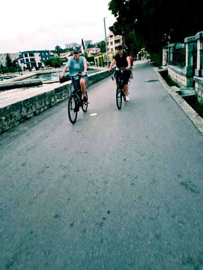 Two people are riding bicycles along a paved path beside a body of water, likely a bay or river, as suggested by the surrounding buildings and vegetation. The path is bordered by a stone wall on the water side and a concrete barrier on the land side, which is lined with trees and buildings. In the background, there are several multi-story buildings with tiled roofs, typical of Mediterranean architecture.
The activity is cycling, with both individuals appearing to be in motion. The person in the foreground, wearing a light blue t-shirt and shorts, is looking towards the camera and has a bicycle with black tires and a visible chain. The person in the background is wearing a black t-shirt and shorts and is also on a bicycle. The lighting suggests it is either early morning or late afternoon, with diffused light and a generally overcast or hazy sky. The overall mood seems relaxed and active, as they are enjoying a bike ride in an urban coastal environment. No visible text is present in the image. The crops suggest that the location is Đenovići, Montenegro.