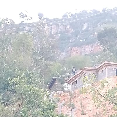 This image captures a landscape scene dominated by a forested hillside and ongoing construction.

In the background, a large, steep hillside rises, densely covered with green trees and bushes. Several prominent sections of exposed rock, appearing light brown or reddish-brown, break through the foliage, indicating the geological structure of the hill. Thin, light-colored lines, possibly power lines or utility cables, stretch horizontally across the upper part of the hill. The sky above is very bright, almost white, suggesting either an overcast day or atmospheric haze.

The midground features a cluster of green trees and, more distinctly, two structures on a sloped, reddish-brown earth surface. To the right, a partially constructed building is visible. It appears to be made of concrete blocks, and the wooden framing for a roof is clearly in place, indicating it is unfinished or undergoing construction. A person, dressed in dark clothing with a light-colored circular emblem or design on their back, stands on the roof or upper level of this building, looking towards the left. To the left and slightly below this main structure, another smaller, darker building or shed, which seems more complete, is partially visible and nestled into the slope, somewhat obscured by trees. A wire or chain-link fence runs along the slope in the lower midground.

The foreground is filled with dense, green foliage and tree branches, some slightly out of focus, framing the bottom and lower sides of the image.

The primary activity depicted is the presence of a person on a building under construction, suggesting either work or observation related to the building project. The overall impression is a developing area set against a natural, mountainous backdrop.

Notable details include the reddish-brown earth indicating exposed soil, possibly from recent excavation or grading for the construction. The time of day appears to be daylight, though the soft, diffused light suggests either an overcast sky, early morning, or late afternoon. The image quality is somewhat soft and grainy.

In the bottom-left corner of the image, the brand watermark "TECNO" is visible.
