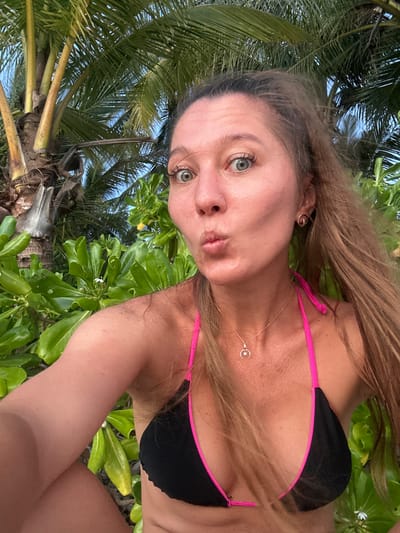 In Baan Taling Ngam, Thailand, a woman is captured in a selfie. She is wearing a black bikini with pink trim and a delicate necklace with a crescent moon pendant. Her hair is long and light brown, and she has green eyes. She is puckering her lips and looking at the camera. Behind her, lush green foliage and palm trees create a tropical backdrop, suggesting a beachside or garden setting. The lighting indicates it is either early morning or late afternoon.
