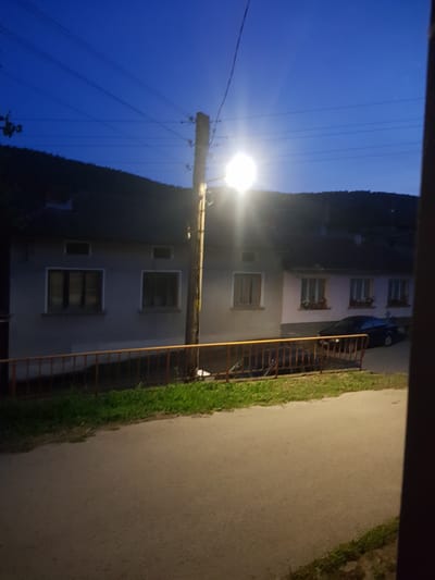 The image captures a street scene in Lomets, Bulgaria, at dusk. A streetlamp illuminates the foreground, casting a bright glow. The street itself is paved and curves towards the left. A grassy area borders the street, with a low orange fence running parallel to it. Behind the fence are several buildings, appearing to be houses, with windows and visible rooftops. A dark-colored car is parked on the street in front of one of the houses. Overhead, power lines stretch across the deep blue twilight sky, which is a characteristic of Lomets. The overall impression is a quiet, residential evening in the Bulgarian countryside.