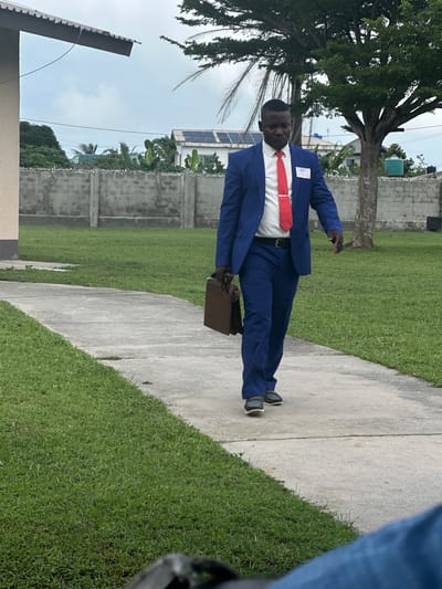 A Black man in a blue suit, white shirt, and red tie is walking on a concrete path outdoors. He is carrying a brown briefcase in his left hand. The path is bordered by green grass on both sides. In the background, there is a grey concrete fence, trees, and buildings with solar panels on their roofs. The sky is overcast. The scene appears to be in a residential or institutional area, possibly in the city of Itoga, Nigeria.