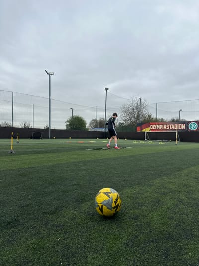 A medium, eye-level, outdoor shot depicts a soccer training session on an artificial turf field. A soccer ball sits in the foreground on the right side of the frame. In the middle ground, a young male figure dressed in dark sportswear is positioned mid-motion, seemingly engaged in a training drill. Behind him, the field is bordered by a black wall with a vertically-oriented banner that reads "OLYMPIASTADION" with a German flag design and logo above the lettering. A tall chain-link fence runs along the edge of the field, partially obscuring a line of trees in the distance. Exercise obstacles can be seen sporadically near the banner. The sky is overcast. The location is identified as Dartford, United Kingdom.