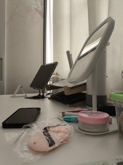 The image shows a white desk with various items, including a smartphone, makeup powder, makeup sponges, a makeup mirror with LED lights, a tablet on a stand, and a keyboard. A light-colored curtain is visible in the background. The overall scene suggests a personal vanity or workspace. The location of General Trias, Philippines is not determinable from the visual information provided in the image.