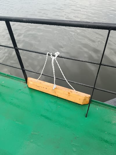 The image shows a portion of a boat deck with a green painted surface. A black metal railing runs horizontally across the scene, with water visible beyond it. Attached to the railing is a piece of light-colored wood, possibly a fender or bumper, secured with a white rope. The wood is positioned horizontally, extending outwards from the boat. The water appears calm with ripples. The scene is likely on a boat docked or moving on the Sava River in Šabac, Serbia, given the context.
