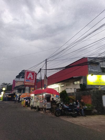 The image displays a street scene in the city of Phước Hòa, Vietnam, under an overcast sky. The environment is a street lined with commercial establishments and vendor stalls. Numerous black electrical wires crisscross the upper portion of the frame, originating from a utility pole near the center-right.
On the left side of the street, a red and white "Alfamidi" sign with a prominent "A" logo is visible, indicating a convenience store. Adjacent to it, a light-colored building with a dark roof is seen. Further right, a large building features a distinct red corrugated roof and light grey walls. A bright yellow rectangular sign, partially visible, reads "RUMAH MAKAN BAKUL SUNDA."
Along the right side of the asphalt road, several street vendor carts are positioned. The leftmost visible cart has a light orange umbrella. Two central carts are covered by red umbrellas; one appears to be a food stall, and a bicycle is parked nearby. A blue motorcycle is parked further right.
At least five individuals are present in the scene. Two individuals are standing near the vendor carts on the far left. One person is standing behind the central red-umbrella vendor cart, possibly attending it. Two individuals are seated on a low wall or curb next to the blue motorcycle on the right, engaged in apparent social interaction. The overall lighting suggests either late afternoon/early evening or a heavily overcast day, with some artificial lights already active.