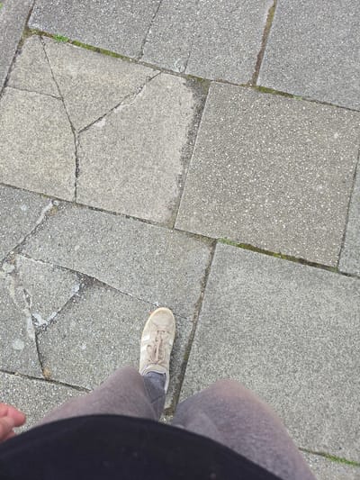 The image shows a person's feet and lower legs as they stand on a cracked concrete sidewalk. The sidewalk is composed of large, square paving slabs, some of which are broken. There are small patches of green grass growing in the cracks between the slabs. The person is wearing a light-colored athletic shoe and grey trousers. The background is entirely composed of the sidewalk. There is no indication of the specific location within Stockton-on-Tees, United Kingdom.