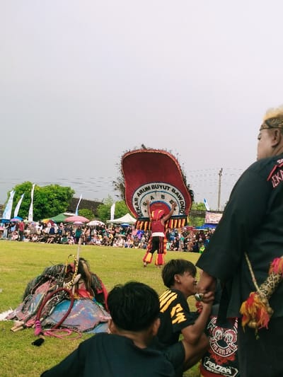 The image was taken in Karangmalang, Indonesia, at the coordinates -7.47 latitude and 111.064 longitude. The scene shows a performance of traditional Indonesian dance. A man in a red and yellow costume is performing a dance with a large, red, feathered headdress. The headdress is decorated with the words "Sekar Arum Buyut Bawono" and features feathers that extend out from the sides of the head. There are people in the background, watching the performance. The performer is holding a staff or cane in his right hand, and he appears to be leaping or spinning. The dancer is surrounded by a crowd of people sitting in a grassy area, watching the performance. Some of the audience members are wearing hats, scarves, and other accessories. The scene appears to be a celebration or festival. The day is partly cloudy, and the ground is covered in a thin layer of grass. To the right of the frame, an older man is wearing a black shirt with a large eagle design.