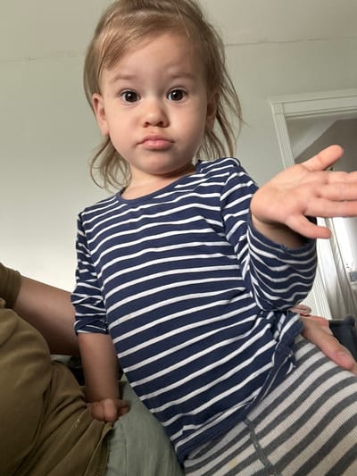A young child is shown from a low angle indoors. The child has blonde hair and brown eyes, and is wearing a navy blue and white striped shirt. The child's right hand is raised with the palm facing the viewer. The child appears to be sitting or lying on a striped surface. Part of an adult's arm and khaki-colored clothing are also visible. The background appears to be a plain wall. The image does not contain any information that could identify the location in Kharnavy, Russia.
