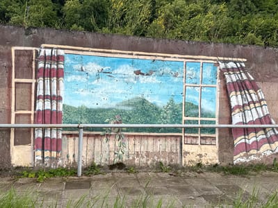 The image displays a mural painted on a concrete wall, depicting an open window with curtains on either side, looking out onto a landscape of rolling green hills under a bright blue sky with scattered clouds. The mural is partially weathered, with some paint peeling and fading. A simple metal railing runs horizontally across the lower portion of the mural, and a few weeds are growing at the base of the wall and on the paved ground in front of it. The wall itself is topped by a dense growth of green trees and bushes, some with small yellow flowers. The scene appears to be in an outdoor urban setting, likely a park or a public walkway, with the concrete paving stones and the railing suggesting a man-made area. The location is identified as Dilijan, Armenia, known for its lush natural beauty.