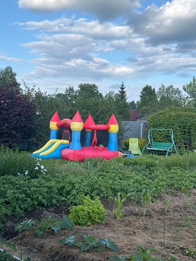 An outdoor, eye-level, medium shot captures a vibrant inflatable castle bounce house situated in a lush, green garden. The bounce house, a colorful structure of red, yellow, and blue, features turrets, a slide, and a net enclosure. It is positioned on a grassy area amidst dense foliage, with trees forming a backdrop. To the right of the bounce house, a green metal swing set with a wooden seat is visible, along with a lime green plastic chair. The foreground is dominated by a vegetable garden, showing rows of leafy plants, including what appear to be potato plants, lettuce, and zucchini or squash. Mulch covers some of the soil. In the distance, a corrugated metal fence and a red-roofed structure can be seen, suggesting a residential or rural setting in the village of Dunilovo, Russia. The sky above is partly cloudy, with large, fluffy white clouds interspersed with patches of blue. The overall scene conveys a sense of outdoor leisure and domesticity.