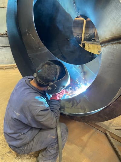 A welder in full protective gear is welding a large, black, spiral metal structure indoors. The welder is wearing a welding mask, gloves, and a blue work suit, and is positioned inside the structure, creating sparks. The environment looks like a workshop or industrial space. A location within the city of Tomba, Montenegro, is not verifiable from this image.