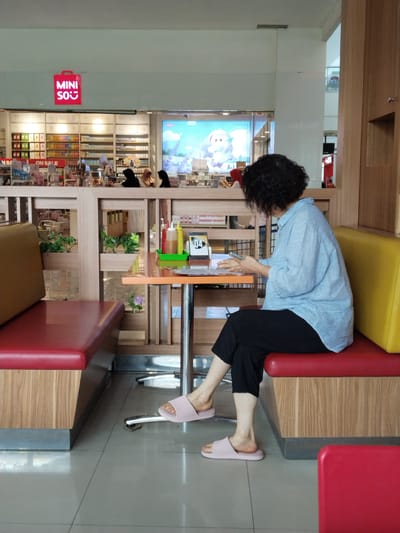 A woman with dark, curly hair, wearing a light blue collared shirt and black pants, sits on a red bench at a table in what appears to be a commercial establishment, possibly a cafe or restaurant. She is looking down at a smartphone or tablet in her hands, which rests on the table. The table is orange and supported by metal legs. On the table are a green tray holding two squeeze bottles of condiments (one red, one yellow) and a glass bottle. The seating area features red and yellow upholstered benches with wooden bases. In the background, shelves stocked with various items are visible, suggesting a retail store. A sign with the word "MINISO" in red lettering and a smiling face is displayed prominently on the wall above the shelves. The floor is tiled in a light grey color. The overall setting is indoor, with bright lighting. The image was taken in Kudus, Indonesia.