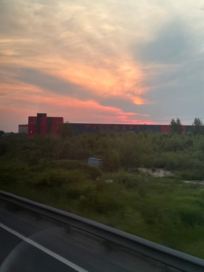 The image depicts a large industrial building in Nikol'skoe, Russia, silhouetted against a sunset sky. The building, painted in a striking combination of red and dark gray, has a prominent red section with multiple windows and a darker section with large lettering that appears to read "РУССКАРТ". A dense line of green trees and bushes separates the foreground from the building. In the foreground, a dark gray barrier with a white stripe, likely a highway guardrail, suggests the perspective is from a moving vehicle. The sky is filled with dramatic clouds, painted in hues of orange, pink, and gray, indicative of twilight. A small blue structure is visible amidst the greenery. There are no people visible in the image.