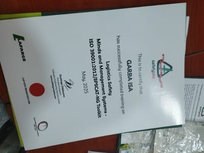 The image is a certificate for a training course on Logistics Safety Management Systems, specifically the ISO 39001:2012/SPSCAT-NG Toolkit. The certificate is issued to GARBA ISA, who has successfully completed the training on an unspecified date. The certificate is issued by Lafarge, a global building materials company, and is dated May 2025. The signature of Damilola Odubunmi, who is presumably a representative of Lafarge, is also present on the certificate. The image appears to have been taken in Ashaka, Nigeria, which is located at the coordinates 10.929, 11.476. The certificate is lying on a wooden table, with other papers visible in the background.