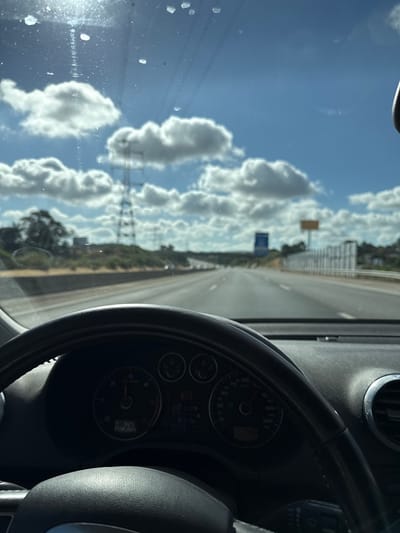 The image is a first-person view from inside a car, looking out the windshield. The scene depicts a highway in Palhais e Coina, Portugal, under a bright, cloudy sky. In the foreground, the top portion of a steering wheel and part of the car's dashboard, including the instrument cluster, are visible. The instrument cluster shows the date as "23.06.2020" and the time as "17:02". The highway stretches into the distance, flanked by trees and vegetation on either side. Power lines and pylons are visible in the background, as well as road signs. The sun is shining, creating some glare on the windshield.