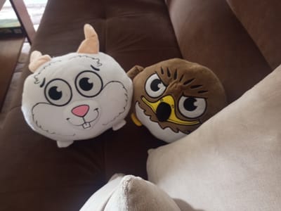 Two plush toys are positioned on a brown sofa. The toy on the left is white and resembles a rabbit or bunny, featuring large black and white eyes, a pink nose, and two light brown horns. The toy on the right is brown and appears to be an owl, with large black and white eyes and dark brown markings around its face. The background consists of the textured brown fabric of the sofa. A cream-colored pillow is partially visible in the foreground, to the right of the plush toys. The overall scene is an indoor setting, likely a living room. There is no visible indication of the city of Radishevo, Bulgaria.