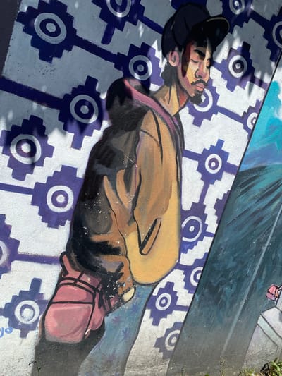 The image shows a painted mural on a surface, likely a sidewalk or pavement. The mural depicts a reclining man with brown skin and dark hair. He appears to be sleeping or resting, and is wearing a dark cap. His clothing is indistinct, but appears to consist of a yellowish-brown garment. His right hand, colored red, is prominently visible, clenched into a fist. The background is patterned with alternating rows of white and blue motifs resembling gears or flowers. The mural is partly obscured by shadows, and portions of an adjacent mural, featuring blue and pink tones, are visible at the top. The image was taken in Quito, Ecuador.