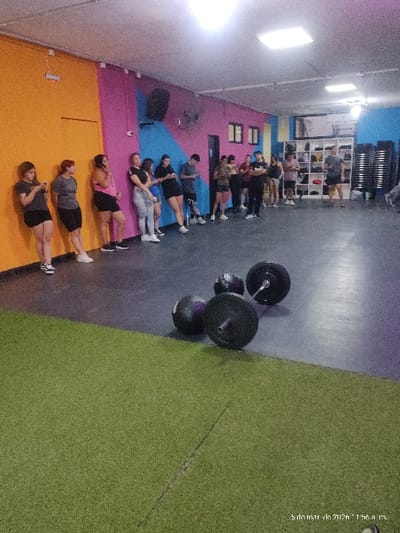 The image displays an indoor fitness facility located in Quilmes, Argentina. Approximately 18 individuals, primarily young adults in athletic attire, are depicted. Most subjects are standing or leaning against a brightly painted wall, which features distinct sections of orange, magenta, and blue. Several individuals are observed looking at mobile phones. In the immediate foreground, a dark gray rubberized floor area contains two barbells fitted with black weight plates and two black medicine balls. This floor transitions into a green artificial turf-like surface further into the foreground. The walls in the background feature a mounted black speaker, a black wall-mounted fan, and two small, dark rectangular windows. To the far right, a white open shelving unit stores various items, and a stack of black chairs or benches is visible. The facility is illuminated by multiple rectangular fluorescent light fixtures on the ceiling. No active exercise is being performed; the individuals appear to be resting or awaiting instruction.