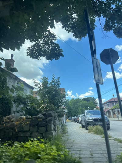 The image shows a view from a moving vehicle on a street in Beloslav, Bulgaria. On the left, there is a stone wall and the facade of a building with a chimney. Trees and foliage are visible throughout the scene, with lush green plants in the foreground and leafy trees lining the street. A sidewalk is present, and several cars are parked along the street. One car, a grey SUV, is prominently featured in the foreground. Traffic signs are visible on poles along the sidewalk. The sky is blue with scattered white clouds. The overall impression is of a residential street in a town on a sunny day.