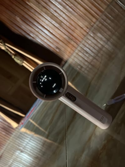 The image shows a close-up, overhead view of a light pink hair dryer lying on a textured surface, possibly a floor or table with a wood-like or tiled pattern. The hair dryer is positioned diagonally, with its nozzle facing upwards and to the left. The nozzle has a black circular insert with white text and indicator lights. The text reads "AIR VOLUME" and "POWER", with several small illuminated dots arranged to indicate different settings. A symbol resembling a gust of wind or heat is also visible. The body of the hair dryer has two small buttons and a subtle embossed "B" logo on the handle. A light purple wrist strap is attached to the end of the handle. The background consists of parallel wooden slats or planks that are darker in color and appear to be part of a piece of furniture or a structural element. The lighting in the scene is dim, creating shadows and highlighting the textures of the surfaces. There are no people visible in the image.

The location in the city of Turumame, Indonesia, cannot be determined from the visual information provided in the image.