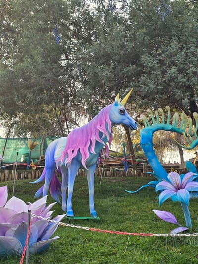 The image depicts an outdoor scene featuring illuminated art installations, possibly as part of a display or festival, which were captured at "Parque Pueblito de Las Vizcachas" in Puente Alto, Chile. There is a large blue unicorn with a pink mane and golden horn is standing on the green grass. To the right of the unicorn is a giant illuminated blue and purple flower display. In the background a green tree is visible. The scene is captured in daylight, resulting in natural lighting. The camera angle is at eye level. The scene appears to be taking place during the day. There is a green fence in the background of the unicorn. There are several chains in the background also.