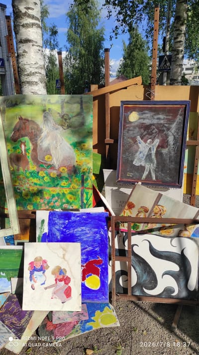 This media file captures an outdoor art display, likely a market or exhibition, in Iisalmi, Finland. The scene is a vibrant, slightly chaotic arrangement of paintings propped up on stands and stacked on the ground. The artworks vary in style and subject matter, including abstract pieces with bold black and white swirls, a painting of a horse amidst sunflowers, a depiction of two figures, and a naive art piece featuring a red bird against a blue background. There are also framed paintings, one of which shows a ghostly figure with a yellow moon.

The setting is outdoors, with lush green trees and a bright blue sky visible in the background, suggesting a sunny day. The time indicated on the image is July 8, 2026, at 18:37, implying it's late afternoon or early evening. The ground is covered in dirt and leaves, reinforcing the outdoor nature of the display. There are also some wooden structures and metal framing holding the artwork.

Visible text elements include partial words and phrases that appear to be from labels or signage associated with the art, such as "NAAMU," "RM 96," "DUKPANN," "LÆRU PL," "Malerduken er festet," and "LEKKETSIAVLE." A watermark at the bottom right corner reads "REDMI NOTE 9 PRO AI QUAD CAMERA." The overall impression is one of a lively and creative artistic gathering set against a natural backdrop.