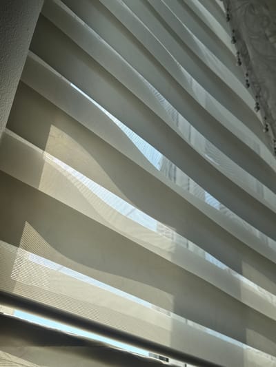 The image shows a close-up view of a window with light filtering through horizontal blinds. The blinds are made of alternating opaque and sheer fabric, creating a layered effect. The light casts shadows and highlights on the blinds, suggesting it is daytime. The texture of the wall to the left of the window is visible, appearing rough and painted. The sheer fabric of the blinds reveals a blurry view of what appears to be a building or structures outside. The scene is indoors, by a window. The location in the city of Benkovski, Bulgaria, is not discernible from the visual information provided in the image. There are no people or actions depicted.