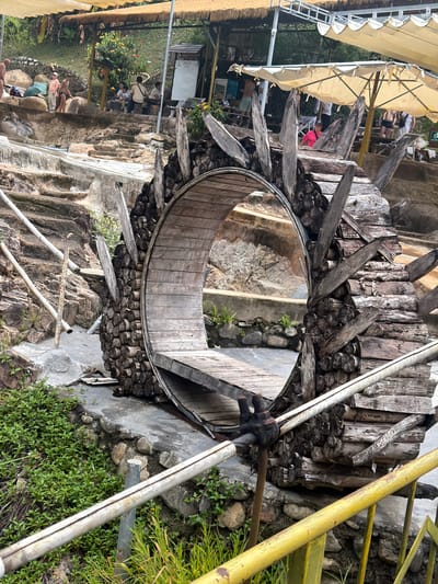 The image depicts an outdoor recreational area, possibly a hot spring or water park, located in the city of Khanh Vinh, Vietnam. In the foreground, a large, rustic wooden structure resembling a wheel or archway is prominently featured. This structure is constructed from logs and planks, with some wooden spikes protruding outwards. A metal railing, partially painted yellow, runs along the bottom of the frame. Behind the wooden structure, a tiered water feature or pools are visible, with stone and concrete steps leading down. In the background, several people are scattered throughout the area, some sitting under thatched and fabric awnings, while others appear to be interacting near the water. The environment is lush with greenery, including trees and bushes. The overall impression is of a natural, somewhat unpolished tourist destination.