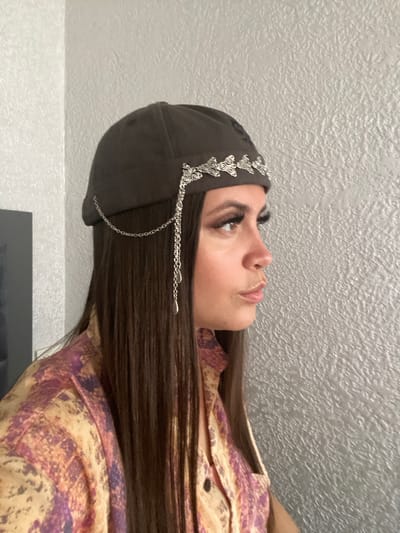 A woman is shown in profile, facing right, against a textured light grey wall. She is wearing a dark brown cap adorned with a silver filigree band and a dangling silver chain with a teardrop pendant. Her long, dark brown hair cascades down her shoulders, partially covering a colorful shirt with a pattern of abstract shapes in shades of yellow, orange, and purple. Her makeup includes prominent false eyelashes and defined eyebrows. The lighting suggests an indoor setting. The location is not identifiable as Chalda, Russia.