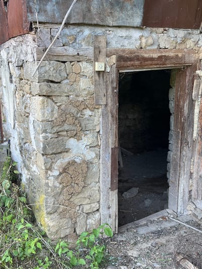 The image captures a ground-level view of an old, deteriorating structure in Byala Reka, Bulgaria, featuring a dark, rectangular opening that appears to lead into an underground space, possibly a cellar or basement. The opening is framed by old, weathered wooden planks. The top plank shows considerable age and wear, with some smaller stones embedded above it, while the bottom wooden beam appears to be bolted into place.
Below the opening, a rugged stone wall is visible, constructed from irregular, light-colored stones cemented with a crude mortar or plaster, much of which is cracked and dried. On the left side of this wall, a faded yellow electrical outlet or light switch is mounted on a small wooden board. To the immediate left of the stone wall, a section of a dark reddish-brown metal sheet or panel can be seen.
The interior of the dark opening is mostly obscured by shadow, but hints of uneven, stony ground are visible, along with a discarded clear plastic bottle and some debris. To the right of the opening, the ground is covered with dry leaves, soil, and some green, leafy vegetation. In the upper right corner, coiled wire and more organic debris are scattered.
No specific activity or event is taking place; the scene is static. The overall impression is one of age, neglect, and disrepair. The natural lighting suggests it is daytime, but no specific time of day is discernible. There is no visible text in the image.