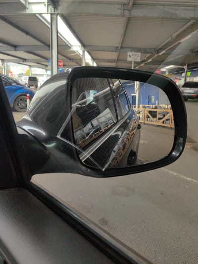 The image depicts a side view mirror of a dark-colored car. The mirror is cracked in the upper left corner. The reflection in the mirror shows the rear of a similar dark-colored car, and a building with blue exterior walls and a fenced outdoor seating area with tables and chairs. Outside of the mirror's view are other parked vehicles, including a blue car, and signs. The location is indicated by the image metadata as Voluyak, Bulgaria. The photograph is taken from inside a vehicle.