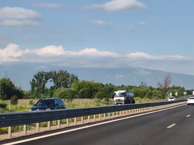 The image shows a highway with several vehicles and a mountainous landscape in the background under a partly cloudy sky. In the foreground, a dark-colored SUV is parked or driving slowly on the shoulder of the road, partially obscured by a guardrail. Further down the highway, a white tanker truck is visible. In the distance, a white sedan is on the highway, and another white vehicle is further ahead. The landscape includes trees and fields. The scene is consistent with a view from a highway, possibly in or near the city of Dolni Bogrov, Bulgaria, given the context.