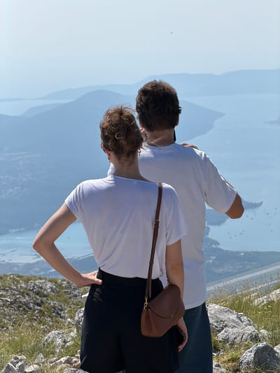 Two people, a man and a woman, stand on a rocky, grassy mountainside overlooking a vast body of water with a hazy, mountainous landscape in the distance. The man, on the left, wears a white t-shirt and dark shorts. The woman, on the right, wears a white t-shirt and black shorts. She has a brown crossbody bag with a brown strap. The image is taken from behind the subjects, looking down a steep incline towards the water. The sky is a clear, pale blue, suggesting a sunny day. The light and shadows indicate it is daytime, likely late morning or early afternoon. The scene conveys a sense of awe and peacefulness, with the couple appearing to be enjoying the panoramic view. The rocky terrain and sparse vegetation are typical of a mountain environment.