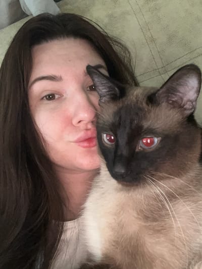 A close-up, selfie-style portrait captures a young woman and a Siamese cat. The woman, with long, dark brown hair framing her face, gazes towards the camera with a slight pout. Her skin appears flushed, and her eyes are brown. Beside her, the cat's face is visible, with its distinctive dark points on its ears and face. The cat's eyes reflect a reddish hue, characteristic of red-eye effect in photography. They are both situated against a textured, olive-green background, possibly a couch or blanket. The image quality suggests it was taken indoors with natural or ambient lighting. There is no indication of the specific location being in the city of Анкудиновка, Russia.