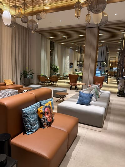 This is an indoor shot of a modern interior, likely a hotel lobby or lounge in the Commune de Diamniadio, Senegal. The foreground is dominated by brown leather sofas adorned with colorful, patterned cushions. Moving towards the back, there's a neutral-toned sofa with an assortment of pillows. Seating is strategically placed throughout, with small tables interspersed.

Architectural elements include light wooden slats on the walls and ceilings, interspersed with white sections. There is a chandelier of glass spheres hanging down from the ceiling near the edge of the image. Tall windows are seen in the distance.

The environment includes indoor plants adding a touch of nature to the modern design. No people are immediately visible, suggesting either a time of quiet or a photograph taken to highlight the design features of the space.