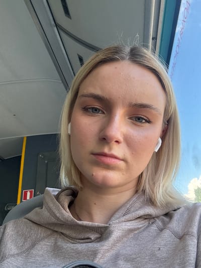 A close-up selfie captured from an upward angle shows a young woman with blonde hair and blue-grey eyes, wearing a light grey hooded sweatshirt. She has a neutral to slightly contemplative expression and is wearing white wireless earbuds in both ears. Her hair falls loosely around her face and shoulders.
The setting appears to be inside public transportation, likely a bus or train, given the overhead compartment-like structures, the window, and the visible signs. Through the window, a bright blue sky with some scattered white clouds is visible, suggesting daytime.
A significant detail is the inverted text visible through the window, which reads "ВЫХОД НА ОСТАНОВКЕ" (VYKHOD NA OSTANOVKE). This Russian phrase translates to "Exit at the stop" or "Stop exit," further indicating the location is a public transit vehicle. Another sign, partially visible in the lower right, shows a red rectangle with a white arrow pointing to the left, commonly used as an exit or direction indicator. The interior features grey panels, a yellow strip, and some dark grey elements.
The crops provide closer views of her face, showing her clear complexion, subtle makeup, and eyebrows, as well as the texture of her sweatshirt and the interior details of the vehicle. The overall impression is a casual portrait taken during travel, possibly in Novoe Devyatkino, Russia.