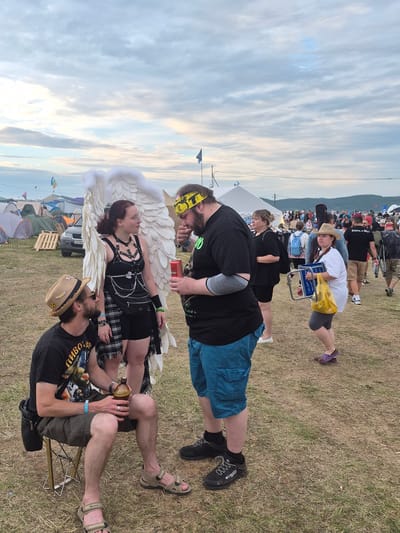 In Lagunovo, Russia, a festival scene unfolds under a cloudy sky. Three individuals are the central focus of the image. On the left, a man wearing a straw hat and sunglasses sits on a portable stool, holding a brown bottle. He is engaged in conversation with a woman standing next to him, who sports large white angel wings, a black top with a harness, and a plaid skirt. The woman is looking towards the man on her right, who is dressed in a black t-shirt and blue shorts. He is holding a red can and appears to be talking to the woman, with his hand near his mouth.
The ground is covered in dry grass and dirt, suggesting a rural or outdoor event setting. In the background, numerous tents are visible, indicating a camping or festival ground. Several other people are scattered throughout the scene, some walking and others standing in groups. A car is parked behind the central figures, and a few wooden pallets are also present. The overall atmosphere suggests a relaxed, outdoor gathering.