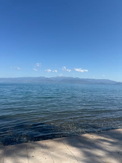 The image shows a clear blue sky with a few white clouds over a calm body of water. In the distance, a mountain range is visible, with some signs of human settlement along its base. The foreground features a paved walkway next to the water, with visible shadows from overhead foliage. The water is shallow near the shore, allowing a view of the pebble-covered seabed. Two small white buoys are floating on the water. The scene appears to be a waterfront area within the Municipal Unit of Kamena Vourla, Greece, likely a public space or promenade. No people are present in the image.