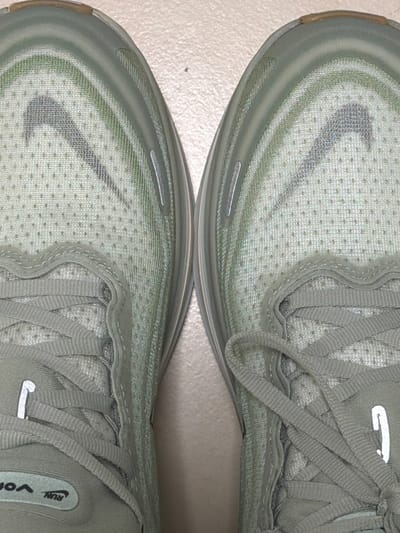 The image shows a close-up of two identical light green Nike running shoes with gray swoosh logos, light green laces, and white reflective tabs on the laces. The shoes are positioned adjacent to each other on a light tan or off-white surface. The texture of the surface appears smooth. There is no identifiable location depicted, and the claim of being in Kfar Saba, Israel, cannot be corroborated from the image. There are no people visible in the image.