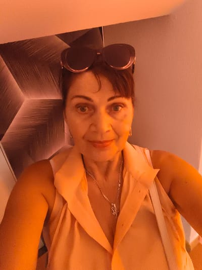 This is a selfie of a woman with dark hair, wearing sunglasses on her head and a light-colored sleeveless shirt. She is also wearing a silver necklace. The background is a wall with a geometric pattern and a plain wall. The lighting suggests it might be indoors, possibly in the evening or under artificial light, as the overall tone of the image is warm and orange-hued. The woman has a neutral expression. There is no visible text. Given the location context of Livorno, Italy, this could be a casual photo taken during a visit or while out and about.