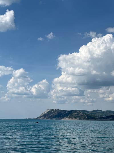 This vertical image, captured in Bol'shoi Utrish, Russia, depicts a coastal landscape under a bright blue sky. The sky, occupying the upper two-thirds, features numerous large, fluffy white cumulus clouds, with some appearing denser towards the right. The lower third of the frame is a calm, turquoise-blue sea with discernible ripples on its surface. Along the distant horizon, a mountainous coastline stretches across the image, characterized by a mix of green vegetation, brown earth, and prominent lighter, possibly rocky, sections. Small, indistinct structures, potentially communication towers or wind turbines, are visible on the highest peaks of the distant land. In the mid-ground of the sea, slightly to the left of the central vertical axis, a dark personal watercraft, likely a jet ski, is observed. A single, dark figure appears to be operating the watercraft, which creates a minor wake as it moves across the water.