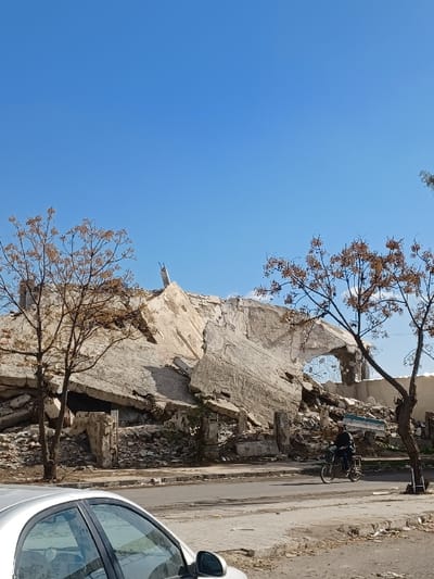 The image depicts an urban street scene in Al-Qadam Municipality, Syria, under a clear blue sky. In the mid-ground, a massive pile of concrete and debris from a collapsed multi-story building occupies the space behind a paved asphalt road and an adjacent concrete sidewalk. Portions of walls and reinforced concrete slabs are visible within the rubble, with a large arched opening discernible on the right side of the demolition site. Two sparse, deciduous trees with brown foliage stand, one on the left and one on the right, partially framing the collapsed structure. On the asphalt road, a solitary individual is observed riding a bicycle or small motorcycle from right to left, wearing dark attire. In the immediate lower-left foreground, the front quarter of a light-colored sedan is partially visible.