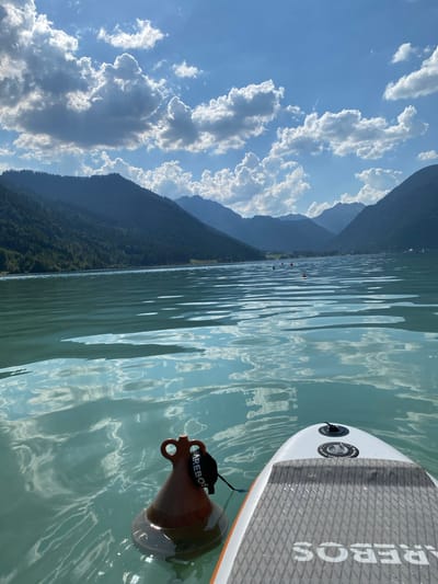 The image depicts a scenic view from a paddleboard on a lake in Lärchenwiese, Austria. The foreground shows the nose of a white paddleboard with grey grip tape and orange trim, partially submerged in clear, turquoise water. The paddleboard has visible text that reads "AREBOS" on the grip tape and a black valve cap with circular branding. Attached to the paddleboard is a reddish-brown anchor with a black strap.

The middle ground features the calm, rippling surface of the lake, reflecting the sky and surrounding landscape. Several small, red buoys are visible in a line across the water, suggesting a designated area or course.

The background is dominated by a majestic mountain range covered in dense green forest, with a hazy silhouette of further mountains in the distance. The sky above is a vibrant blue, dotted with fluffy white clouds, some of which are illuminated by the sun. The lighting suggests it is daytime, likely mid-morning or afternoon, with bright sunshine casting reflections on the water. The overall atmosphere is serene and idyllic, conveying a sense of outdoor recreation and peaceful enjoyment of nature.