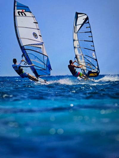 The image shows two windsurfers performing tricks. The water is churning behind them, indicating speed. The background is a clear, blue sky. The upper windsurfer wears colorful shorts, while the lower one sports a white shirt with a "3" on it and dark shorts. The sails of the windsurfers are blue and white. The upper sail has a yellow and black section, while the lower sail has black stripes. The location is Bacong, Philippines.