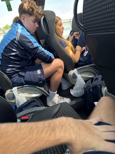 The image captures the interior of a passenger coach, identified as being in Bolsover, United Kingdom. Three passengers are prominently featured, viewed from a forward-facing perspective. On the left, a male, appearing to be a teenager, with light brown, curly hair, wears a navy blue tracksuit jacket with light blue shoulder panels and navy blue shorts bearing a white Canterbury logo. He is seated, looking downward. His lower legs are visible, clad in white athletic socks and white shoes. Adjacent to him, on the right, a female, also appearing to be a teenager, with long, straight light brown hair, wears a dark blue t-shirt. She holds a smartphone in her right hand, which has red-painted fingernails, and wears multiple bracelets on her left wrist. Her mouth is slightly agape, and she is looking towards the right side of the frame. The immediate foreground shows the hairy forearm and upper leg of an unseen individual, presumably the photographer. The coach interior features grey and black patterned seating with grey plastic elements, including footrests or storage areas. A navy blue bag and two white plastic bags are positioned on the floor. An emergency exit sign is partially visible in the upper left corner of the bus window.