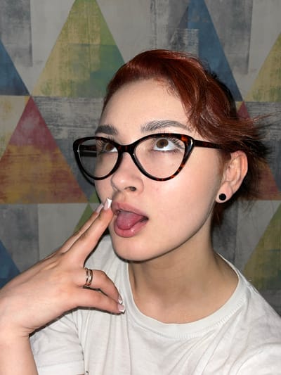 In the city of Starobaltachevo, Russia, a close-up portrait shows a young woman with reddish-brown hair, wearing glasses with tortoiseshell frames. She is looking upwards and to her left, with her mouth slightly open and her tongue visible. Her right index finger is touching her lips. She is wearing a white t-shirt and a silver ring on her left hand. The background is a wall with a geometric pattern of triangles in various colors, including blue, green, red, and yellow. The lighting is artificial and casts soft shadows.