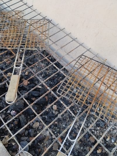 This image displays an outdoor charcoal grilling area located in Lo Ferro, Spain. The scene is dominated by a bed of spent and cooled charcoal pieces, ash, and small stones, covering the ground. A large, square-grid metal grate, exhibiting significant rust and white ash residue, is positioned over this charcoal bed. Resting on this primary grate are two smaller, portable wire barbecue baskets. The basket on the left features a light-colored, rectangular wooden handle and shows areas of rust on its wire mesh. The basket on the right is heavily rusted, particularly on its upper section, and has a U-shaped metal wire handle. A light-colored, textured wall, consistent with concrete or stucco, occupies the upper right background, forming a corner with the ground where the grilling setup is situated. No individuals are present.
