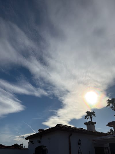 The image displays a vibrant daytime sky over Mont-roig del Camp, Spain, featuring architectural elements and natural phenomena. The primary subject is the bright sun, which is surrounded by a prominent, colorful solar halo, indicating the presence of ice crystals in the atmosphere. The sky is a mix of deep blue patches and dynamic white clouds, ranging from thick and diffuse to wispy, streaky formations.
To the right side of the frame, a segment of a building with white stucco walls and a reddish-brown tiled roof is visible. Its architecture, including a decorative outdoor light fixture, a dark drainpipe, and a window with security bars, evokes a Mediterranean or Spanish style. Several dark silhouettes of palm trees are scattered across the upper portion of the frame, some appearing near the sun and others above the building. A distinct chimney with an ornate cap rises from the roofline. Below the main roofline, an antenna or aerial is visible on a lower section of the roof. A faint white contrail from an aircraft can be seen cutting across a patch of blue sky. The scene suggests a sunny, pleasant day. No text is visible in the image.